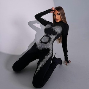 May include: A black jumpsuit with long sleeves and a high neckline. The jumpsuit is embellished with a pattern of sparkling rhinestones. The model is wearing black high-heeled boots.