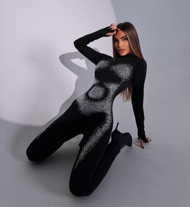 May include: A black jumpsuit with long sleeves and a high neckline. The jumpsuit is embellished with a pattern of sparkling rhinestones. The model is wearing black high-heeled boots.