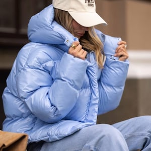 May include: A light blue puffer jacket with a hood and a front zip fastening, worn with light blue denim jeans and a cream baseball cap featuring the text PARIS FRANCE embroidered in dark brown lettering on the front panel.