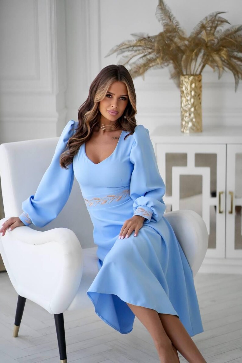 May include: A light blue dress with long sleeves and a V-neck. The dress features a fitted waist with embroidered detailing and a flowing skirt. The sleeves have a slight puff at the shoulder and are gathered at the wrist. The dress is worn while seated in a white chair.