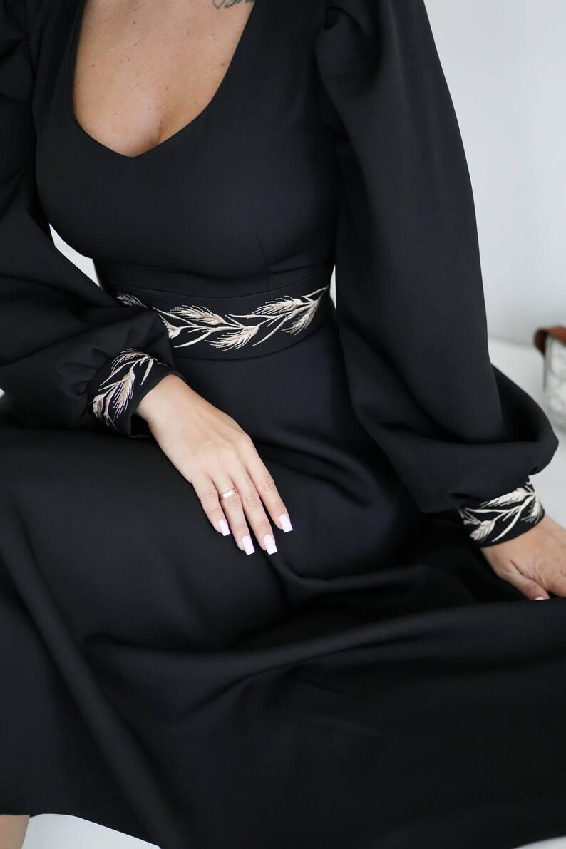 May include: Black dress with a V-neckline and long, voluminous sleeves. The dress features a decorative embroidered design around the waist and cuffs. The dress is a solid black color.