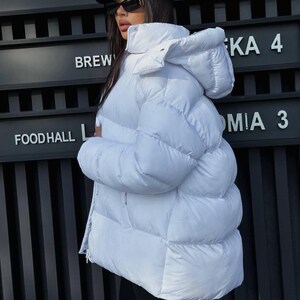 May include: A bright white puffer jacket featuring a high collar, a detachable hood, and horizontal quilted stitching. The jacket has a full-length front zipper closure and a thick, insulated design, providing a voluminous silhouette with long sleeves.
