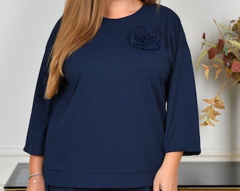 May include: A dark navy blue long-sleeved top featuring a round neckline and a matching fabric flower embellishment on the upper left chest. The garment has a straight hemline and a relaxed fit, crafted from a solid-coloured knit fabric.