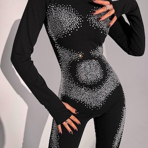 May include: Black jumpsuit with long sleeves and a high neckline. The jumpsuit is embellished with numerous silver rhinestones arranged in a decorative pattern across the bodice and legs. The outfit is form-fitting.