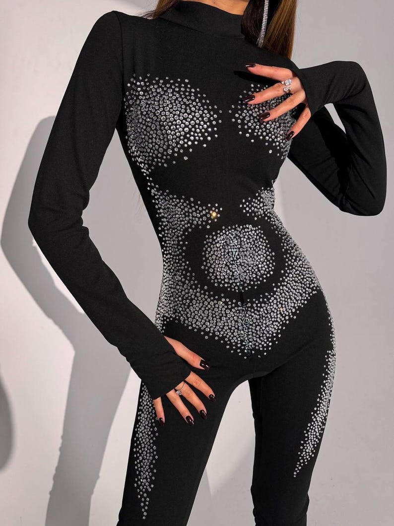 May include: Black jumpsuit with long sleeves and a high neckline. The jumpsuit is embellished with numerous silver rhinestones arranged in a decorative pattern across the bodice and legs. The outfit is form-fitting.