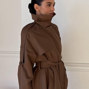 Könnte beinhalten: Ein dunkelbrauner Trenchcoat mit hohem Kragen, Schulterklappen und einem Bindegürtel in der Taille. Der Mantel verfügt über lange Ärmel mit verstellbaren Schnallenriemen an den Manschetten, eine Sturmklappe auf der Brust und tiefe Seitentaschen.