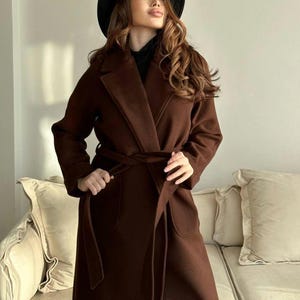 May include: A long, dark brown coat featuring a wide lapel collar, long sleeves, and a matching fabric tie belt at the waist. The coat includes two large front pockets and is worn with a black hat and a black high-neck top underneath.