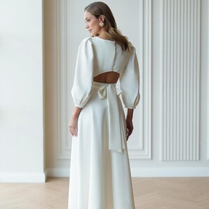 May include: A floor-length white dress featuring long puff sleeves, a square neckline, and a cutout back detail. The garment includes a wide fabric tie at the waist that hangs down the back, creating a structured silhouette with a flowing skirt.