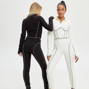 Op de afbeelding: Een zwarte set met een top met lange mouwen en een legging naast een witte set. Beide outfits zijn voorzien van een kraag met kwartritssluiting, duimgaten en opvallende grijze contraststiksels die een korsetachtig lijfje accentueren.