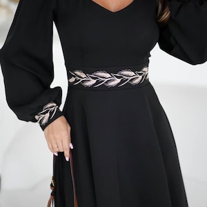 May include: Black dress with long sleeves and a V-neckline. The dress features a decorative embroidered belt and cuffs with a wheat-like design. The dress has a flowing skirt and is suitable for formal occasions.