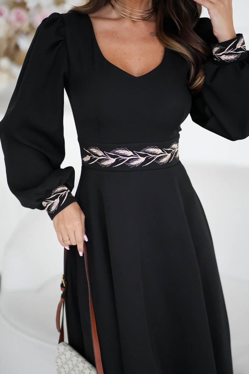 May include: Black dress with long sleeves and a V-neckline. The dress features a decorative embroidered belt and cuffs with a wheat-like design. The dress has a flowing skirt and is suitable for formal occasions.