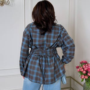 May include: A long-sleeved button-down shirt featuring a brown and blue plaid pattern. The garment includes a corset-style lace-up detail at the waist with matching fabric ties that hang down the back. The shirt is paired with light blue denim jeans.