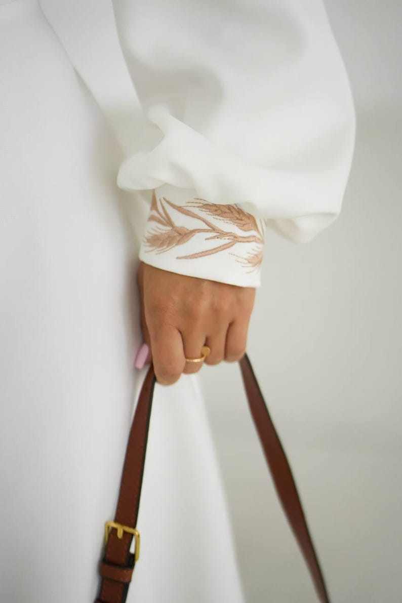 May include: White long-sleeved dress with wheat-like embroidery on the cuff. A brown leather handbag with a gold buckle is held in the hand. The ring on the finger is gold.