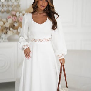 May include: A white dress with long, puffy sleeves and a scoop neckline. The dress features a decorative band at the waist and cuffs. A small, patterned purse with a brown strap is held in the hand. The dress is a midi length.