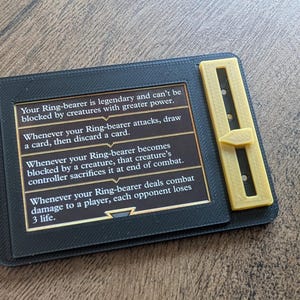Peut inclure: Un porte-carte rectangulaire en plastique noir avec un curseur jaune sur le côté droit. La carte affiche du texte détaillant les règles de jeu pour un porteur d'anneau, incluant des capacités liées au blocage, au tirage de cartes et aux points de vie.