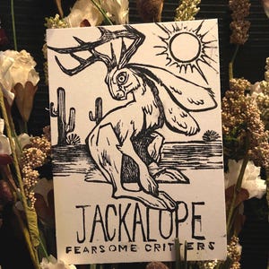 Puede incluir: Un grabado en linóleo en blanco y negro que representa un jackalope con cuernos, un paisaje desértico y un sol. El texto "JACKALOPE" y "FEARSOME CRITTERS" está impreso debajo de la imagen. La impresión está rodeada de flores secas.