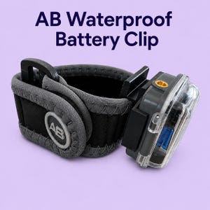 May include: A black and grey waterproof battery clip, with the text "AB Waterproof Battery Clip". It features a clear plastic compartment and a strap with a buckle. The clip is designed for secure attachment.