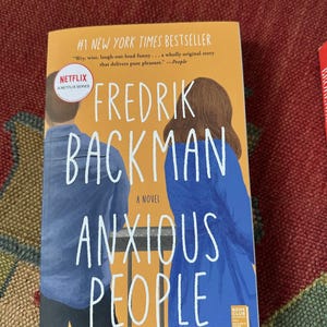 May include: A hardcover book titled "Anxious People" by Fredrik Backman. The cover is orange with the title in large white letters. It is a New York Times bestseller and a Netflix series.