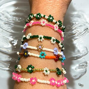May include: Close-up of several beaded bracelets in various colors, including green, pink, white, blue, and gold. The bracelets feature floral designs and are worn on a wrist. The bracelets are made of small beads.
