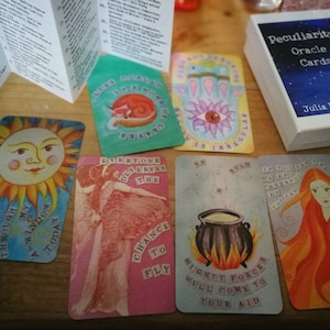 May include: An oracle card set featuring illustrated cards with images of a sun, a fox, a hand, and a cauldron. The deck is called "Peculiarity Oracle Cards" by Julia Breinholt, with a white box and a blue watercolour design.