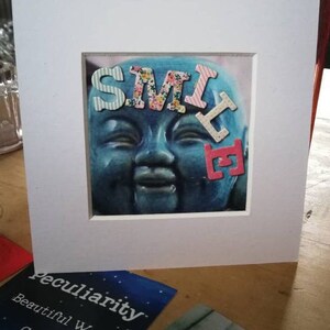 May include: A framed print of a blue Buddha-like face with the word "SMILE" in patterned letters. The print is matted in white. The image is on a wooden surface, with "Peculiarity Beautiful Word Cards" visible in the foreground.