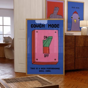 May include: A framed poster with the words "COUCH MODE" above a large pink and green switch, with "ON" and "OFF" labels. Below, it reads "THIS IS A HIGH PERFORMANCE CHILL ZONE." Two other framed art pieces are visible, one with a bird and the other with the text "NICE BUM."