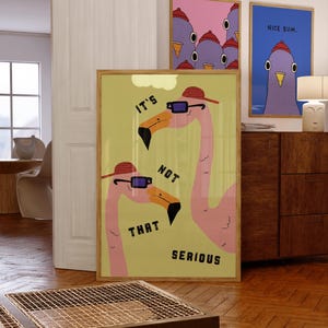 Op de afbeelding: Een ingelijste kunstprint met twee illustraties van roze flamingo's met zonnebrillen en hoeden, met de tekst "IT'S NOT THAT SERIOUS" in het zwart. Twee extra ingelijste prints met vogelillustraties zijn zichtbaar.