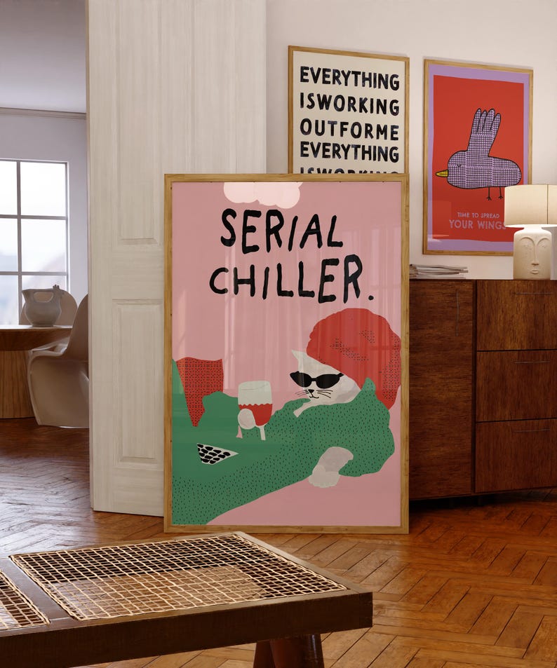 May include: A framed pink poster with the text "SERIAL CHILLER" features a cat wearing sunglasses and holding a glass of wine. The cat is lounging on a green blanket. Two other framed posters are visible in the background.
