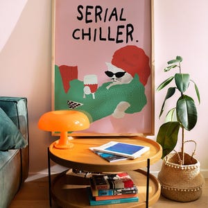 May include: A framed poster with the text "SERIAL CHILLER" above a cartoon cat wearing sunglasses and a red hat, holding a glass of wine. The poster is above a two-tiered wooden coffee table with an orange lamp and books. A potted plant is to the right.