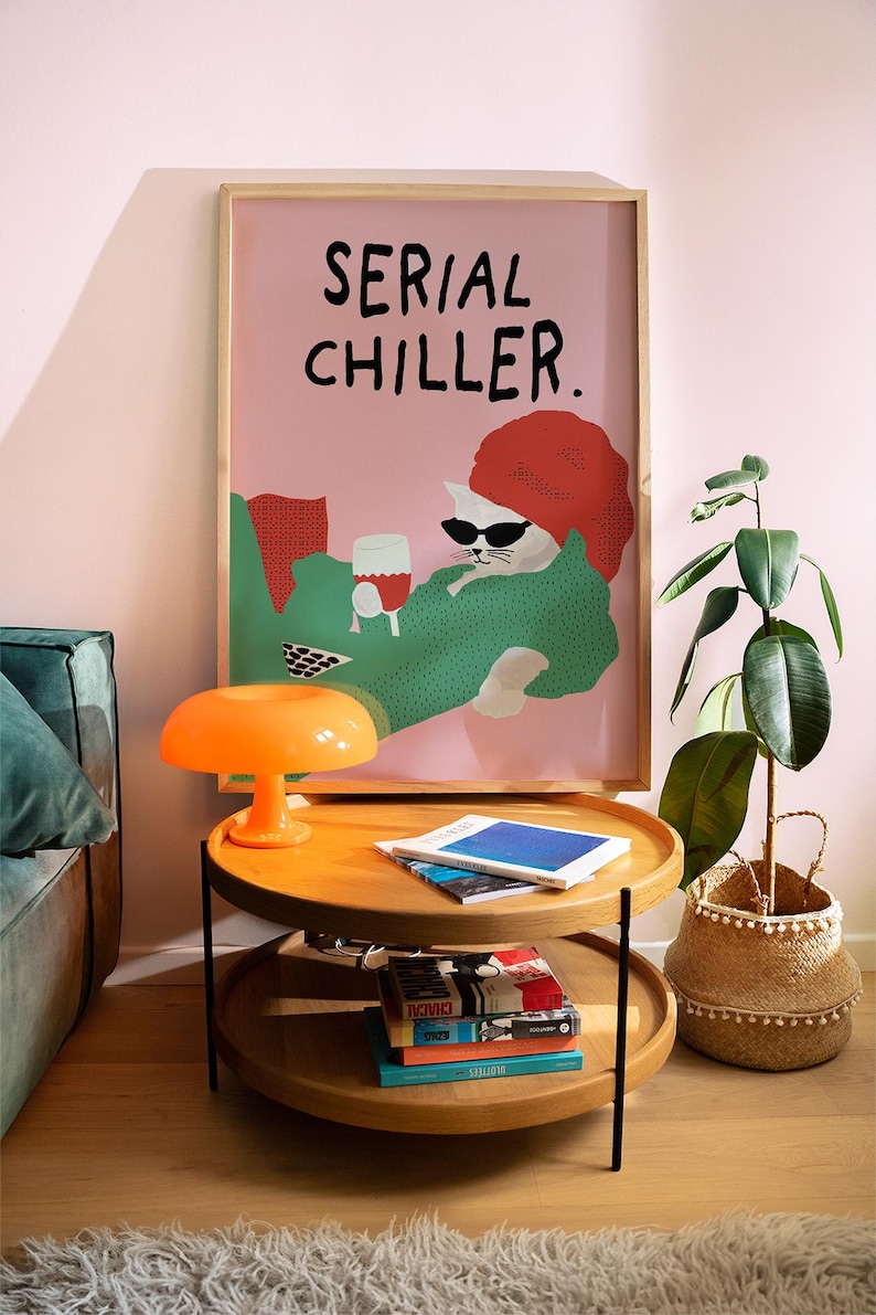 May include: A framed poster with the text "SERIAL CHILLER" above a cartoon cat wearing sunglasses and a red hat, holding a glass of wine. The poster is above a two-tiered wooden coffee table with an orange lamp and books. A potted plant is to the right.
