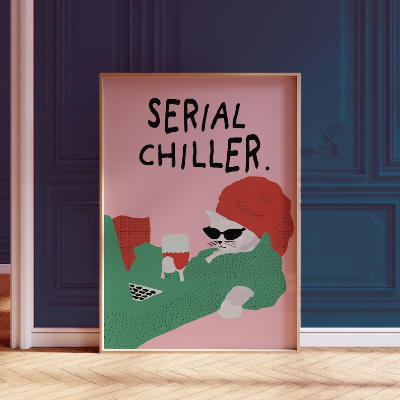 May include: A framed art print featuring a cat wearing sunglasses and a red hat, holding a glass of wine. The text "SERIAL CHILLER" is displayed above the cat on a pink background. The cat is wearing a green sweater.