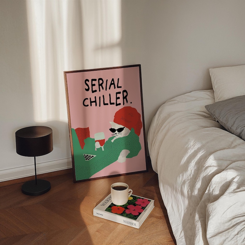May include: A framed pink poster with the text "SERIAL CHILLER" features a cartoon cat wearing sunglasses and a red hat, lounging with a drink. A black lamp, a coffee cup, and a book with floral art are on the wooden floor.