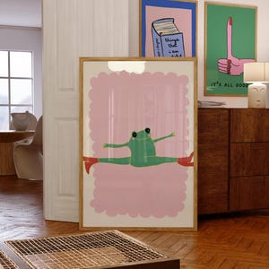 Op de afbeelding: Een ingelijste kunstprint met een groene kikker in spagaat, met rode laarzen, tegen een roze achtergrond met een geschulpte rand. Twee andere ingelijste prints zijn zichtbaar, een met een boek en tekst, de andere met een hand.