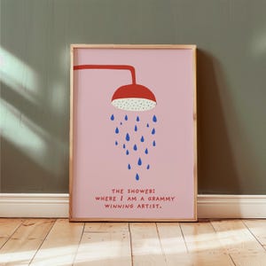 Op de afbeelding: Ingelijste kunstprint met een rode douchekop en blauwe waterdruppels op een roze achtergrond. De tekst luidt: "THE SHOWER: WHERE I AM A GRAMMY WINNING ARTIST."