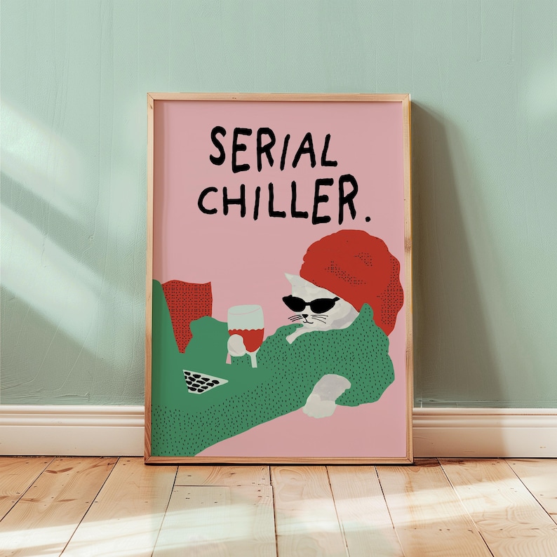 May include: A framed art print featuring a white cat wearing sunglasses and a red hat, lounging with a glass of red wine. The cat is draped in a green blanket, with the words "SERIAL CHILLER" in black text on a pink background.