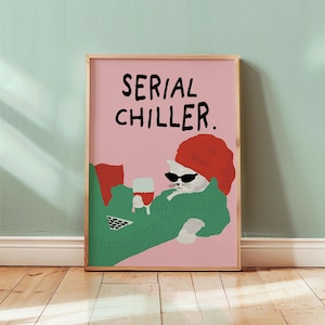 May include: A framed art print featuring a white cat wearing sunglasses and a red hat, lounging with a glass of red wine. The cat is draped in a green blanket, with the words "SERIAL CHILLER" in black text on a pink background.