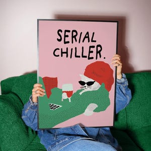 May include: A framed art print featuring a cartoon cat wearing sunglasses and a red beret, holding a glass of wine. The print has the words "SERIAL CHILLER" in black text on a pink background. The cat is wearing a green sweater.