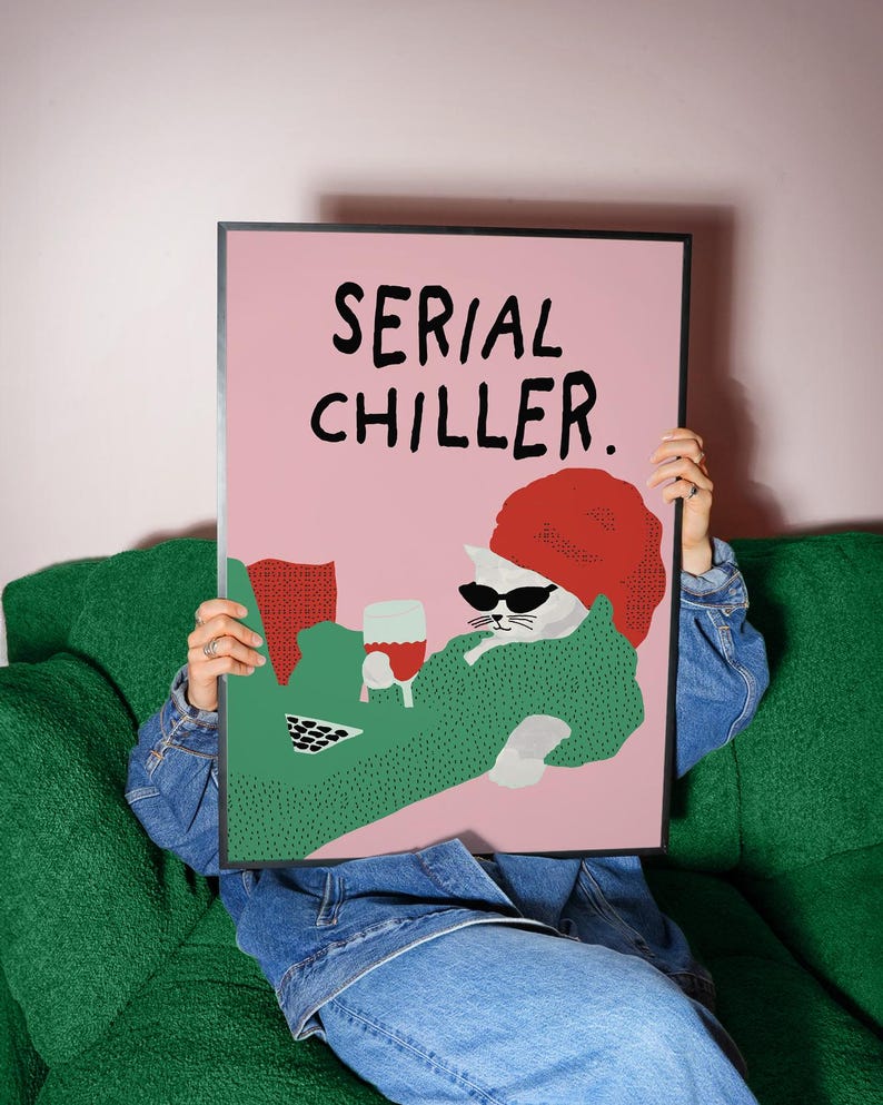 May include: A framed art print featuring a cartoon cat wearing sunglasses and a red beret, holding a glass of wine. The print has the words "SERIAL CHILLER" in black text on a pink background. The cat is wearing a green sweater.