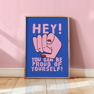 Könnte beinhalten: Ein gerahmter Kunstdruck mit blauem Hintergrund und rosa Text, der lautet: "HEY! YOU CAN BE PROUD OF YOURSELF!". Eine rosa Faust ist in der Mitte abgebildet. Der Rahmen ist hellbraun.