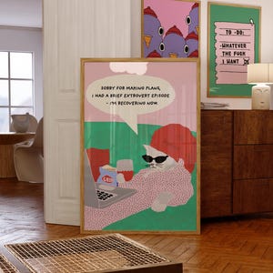 May include: A framed art print shows a cat in sunglasses and a red beanie, using a laptop with snacks. The print includes the text: "SORRY FOR MAKING PLANS, I HAD A BRIEF EXTROVERT EPISODE - I'M RECOVERING NOW."