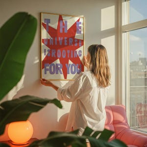 Pode incluir: Uma mulher est&aacute; pendurando um p&ocirc;ster emoldurado em uma parede branca. O p&ocirc;ster tem uma estrela vermelha em forma de explos&atilde;o com o texto "THE UNIVERSE IS ROOTING FOR YOU" em letras roxas.