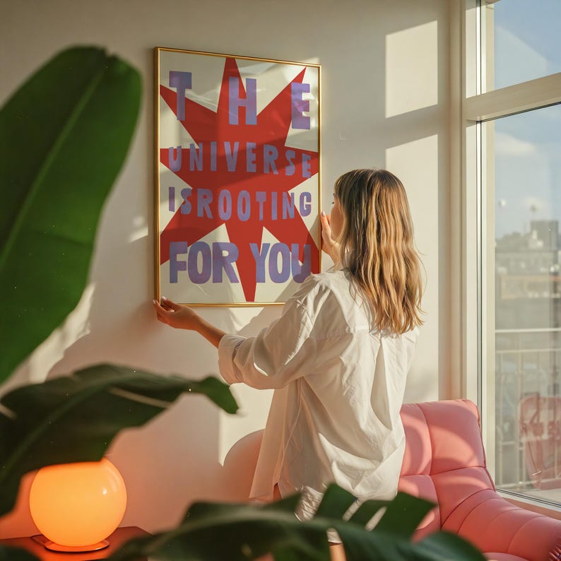 Pode incluir: Uma mulher est&aacute; pendurando um p&ocirc;ster emoldurado em uma parede branca. O p&ocirc;ster tem uma estrela vermelha em forma de explos&atilde;o com o texto "THE UNIVERSE IS ROOTING FOR YOU" em letras roxas.
