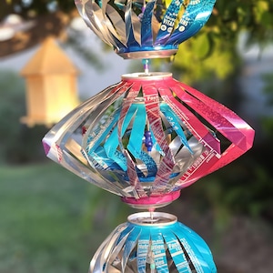 Könnte beinhalten: Ein hängendes Windspiel aus recycelten Aluminium-Getränkedosen, bestehend aus drei gestapelten, kugelförmigen Abschnitten. Die Metallstreifen sind gebogen und zeigen leuchtende Blau-, Pink- und Silbertöne mit sichtbaren Nährwertangaben auf dem Material.
