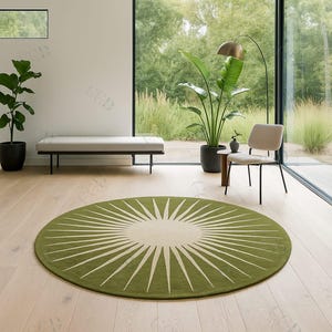 Op de afbeelding: Een ronde, olijfgroene vloerkleed met een crèmekleurig midden en witte, uitstralende lijnen. Het kleed ligt op een lichte houten vloer in een moderne woonruimte. De kamer heeft grote ramen en kamerplanten.