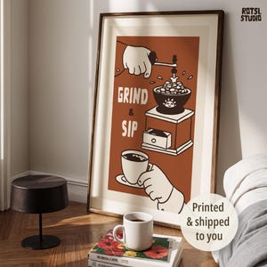 May include: A framed vertical wall art print featuring a rust-coloured background with a graphic illustration of a hand operating a coffee grinder and another hand holding a cup of coffee. The text GRIND & SIP is printed in white capital letters.