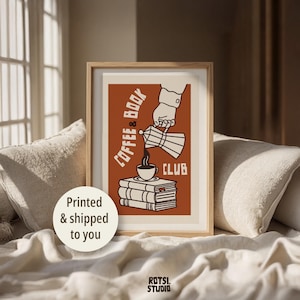 Op de afbeelding: Een ingelijste wandprint met een roestkleurige achtergrond en een grafische illustratie van een hand die koffie uit een percolator in een kopje op een stapel boeken schenkt. De tekst luidt Coffee & Book Club in een crèmekleurig lettertype.