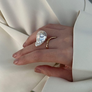 May include: A large, irregularly shaped white pearl ring paired with a delicate gold band adorned with small, sparkling accents. The ring is set against a cream-colored blazer, showcasing the jewelry's elegant design.