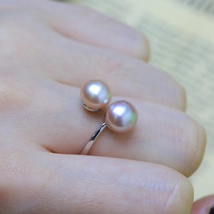 Puede incluir: Un anillo de estilo abierto que presenta dos perlas de color rosa pálido y lustroso montadas sobre una banda de tono plateado pulido. Las perlas se sitúan en cada extremo del aro abierto, creando un diseño equilibrado sobre el dedo.