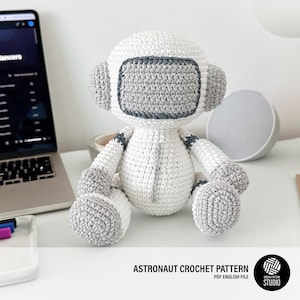 May include: A handmade white and gray crochet astronaut doll. The doll has a round head with a gray visor and ear coverings, and gray accents on the arms and legs. The text "ASTRONAUT CROCHET PATTERN" is visible.