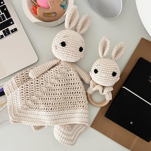 Puede incluir: Dos juguetes de conejo de ganchillo en color crema. Uno es una manta de seguridad grande con una cabeza de conejo, el otro es un sonajero de conejo más pequeño con un anillo de madera. Un cuaderno negro con el texto "NOTES FOR YOUR LIFE" también está en la imagen.